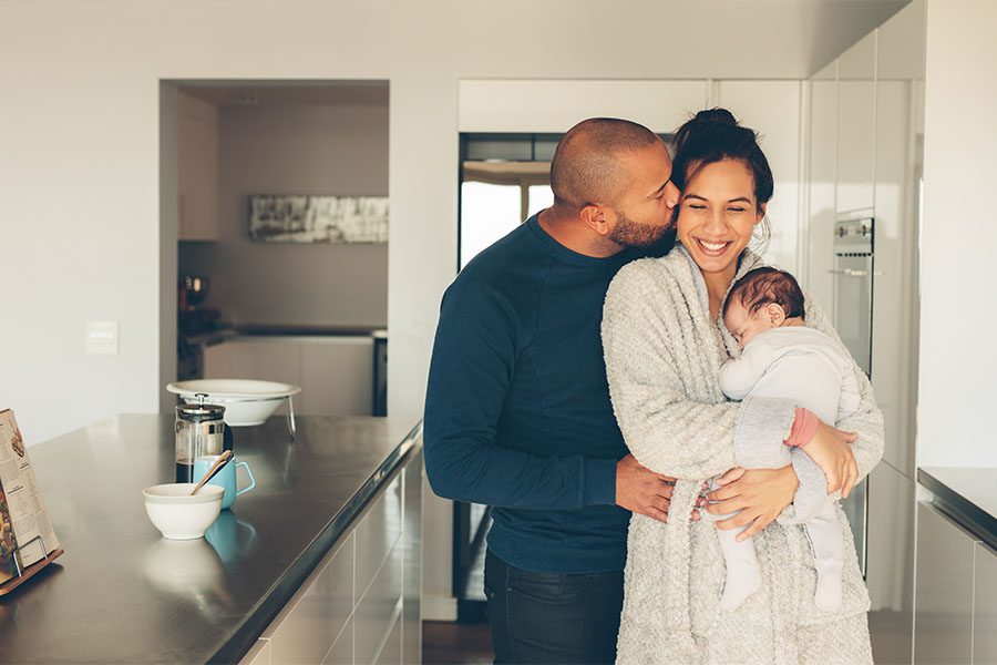 Personal Insurance - Portrait of a Father Kissing His Happy Wife Who is Holding Their Newborn Baby While They Stand in the Kitchen